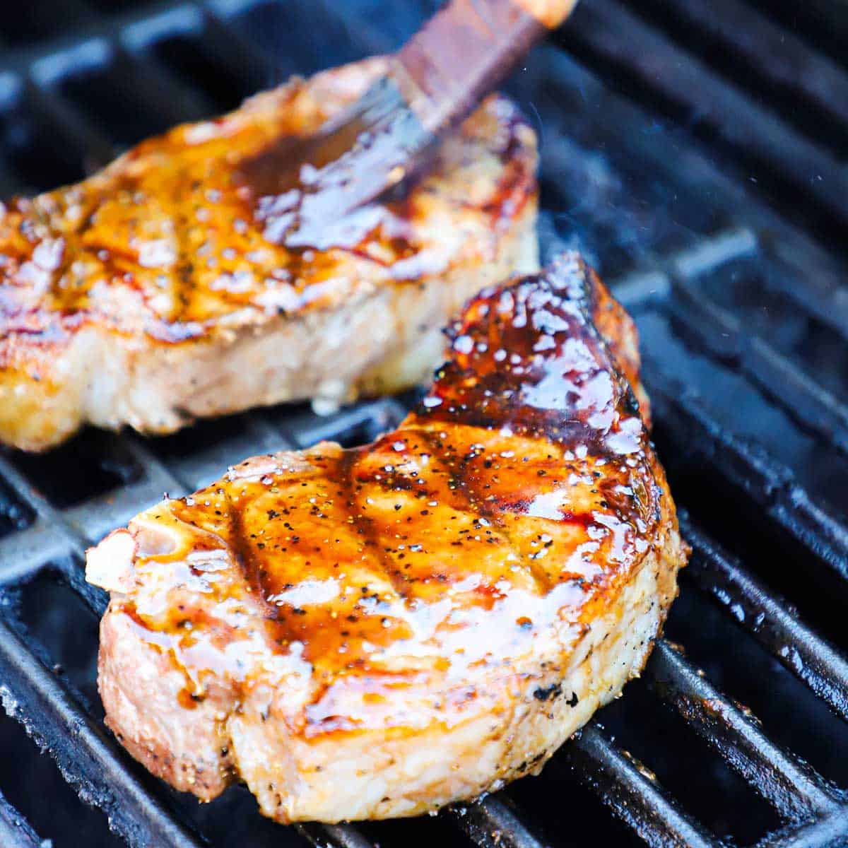 A person using a small brush to baste two pork chops with agrodolce sauce on a gas grill. 