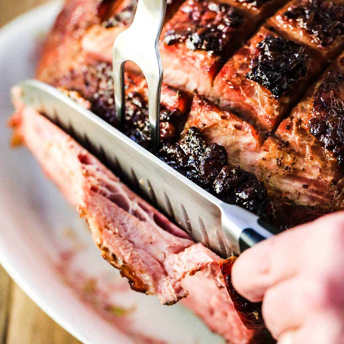 A person using a meat fork and a large carving knife to cut a large roasted ham into slices on a white platter. 