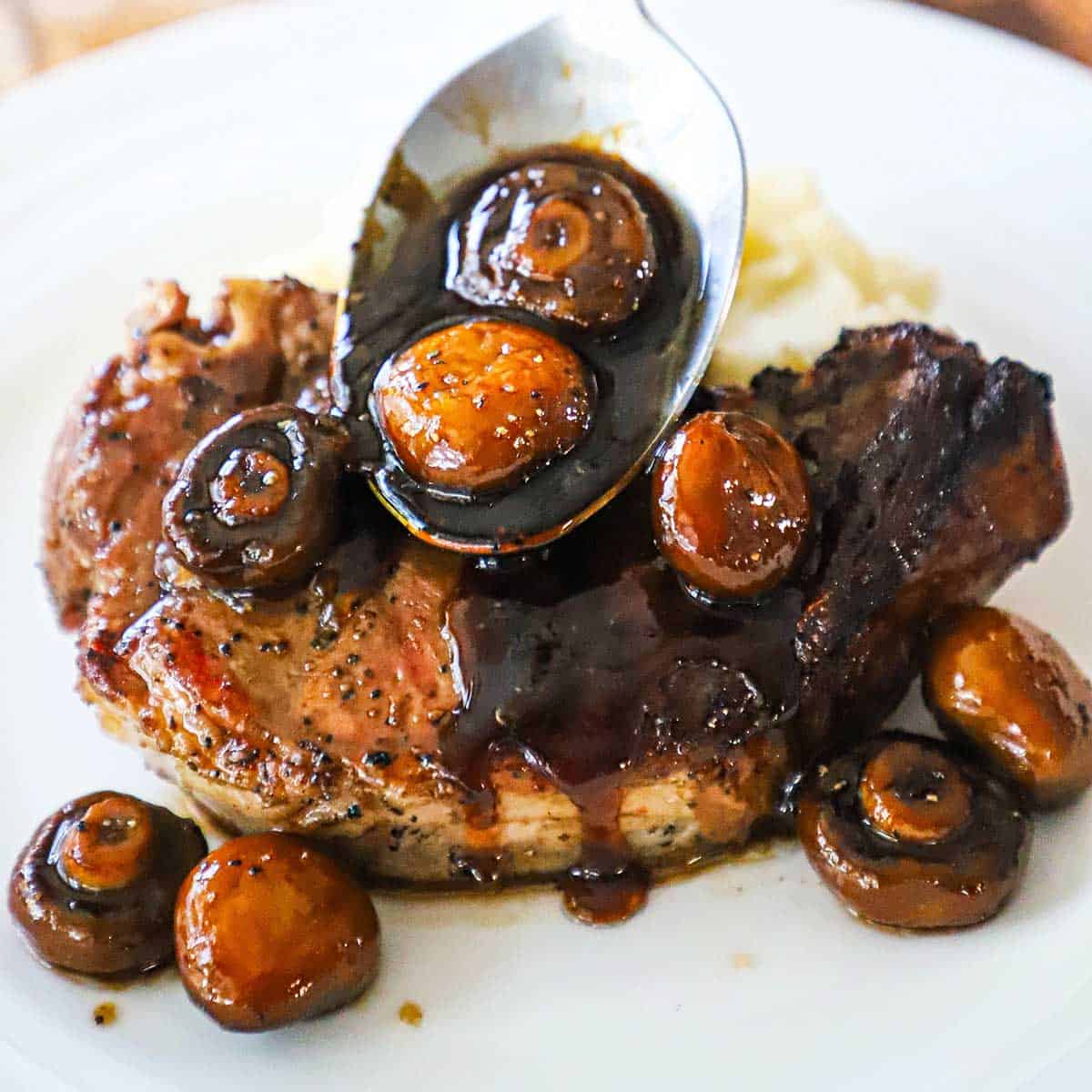 A person spooning an agrodolce sauce with mushrooms over a grilled pork chop on a white dinner plate. 
