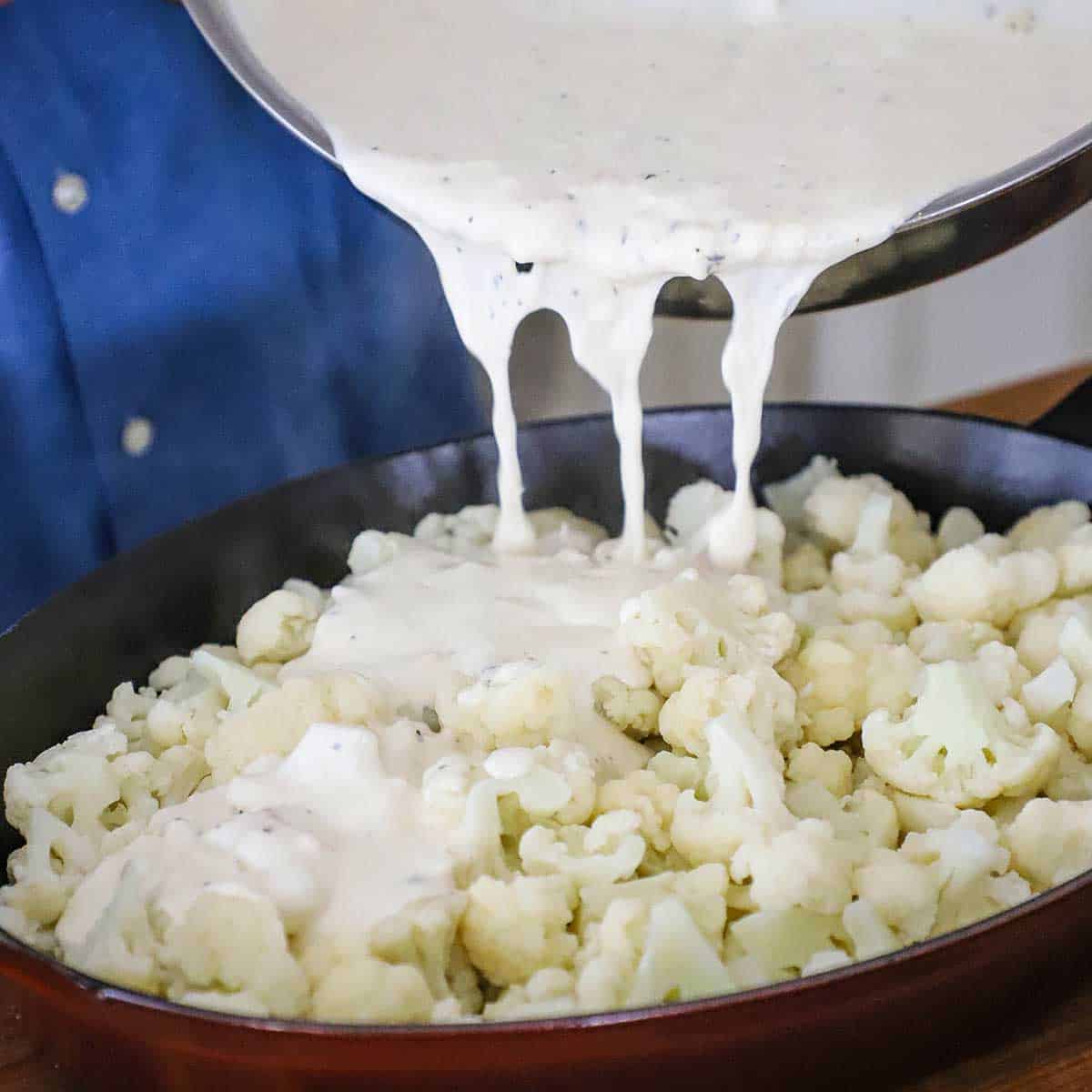 A person pouring a creamy cheesy sauce over cauliflower florets that have been arranged in an oval baking dish. 