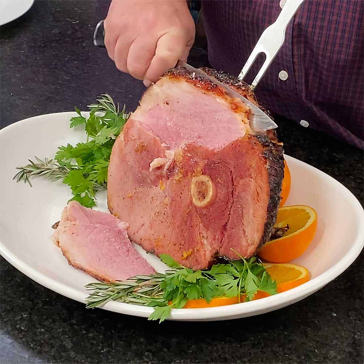 A person using a carving fork and knife to slice into a bourbon orange glazed ham on a white platter. 