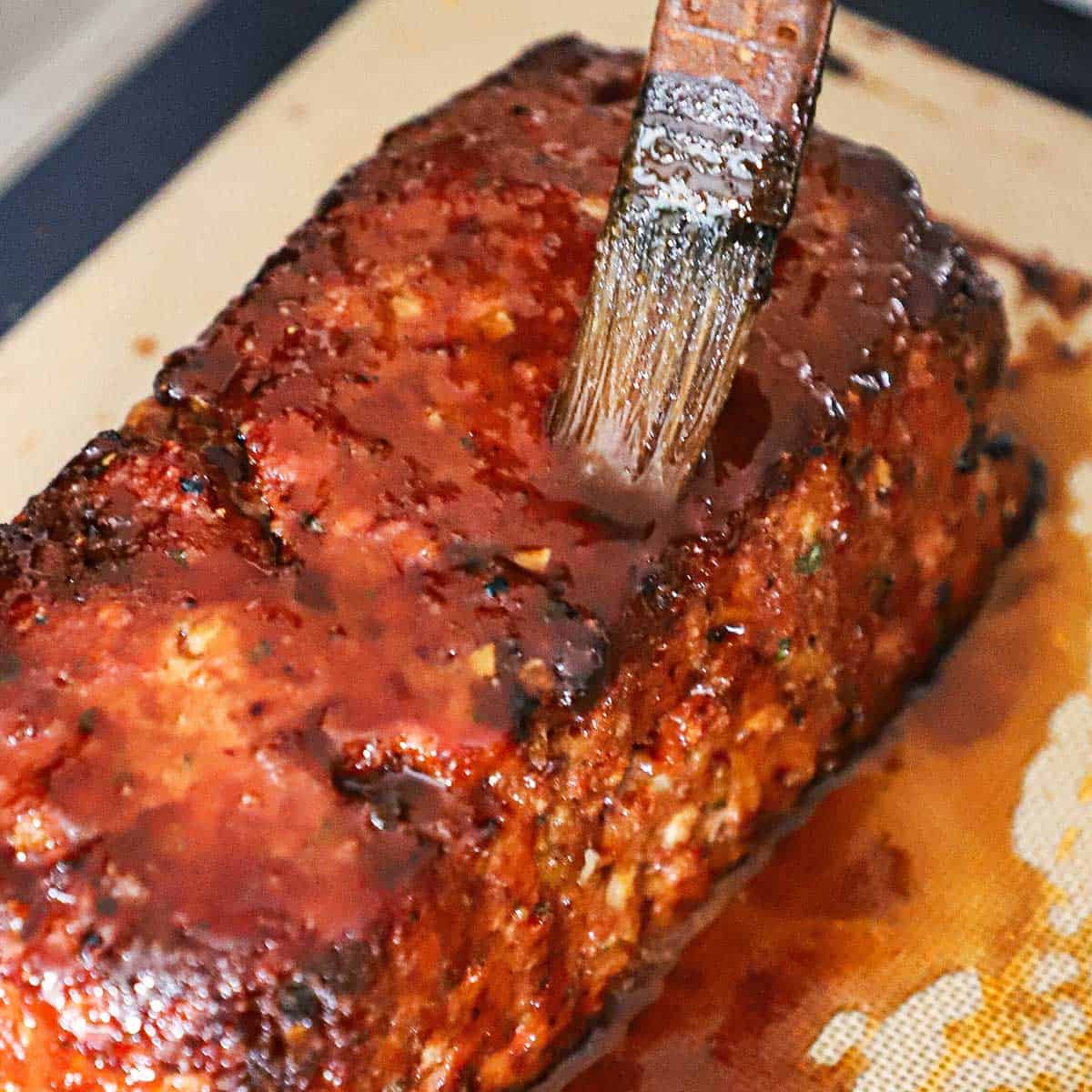 A person using a pastry brush to apply a glaze to a baked ham loaf on a baking sheet. 