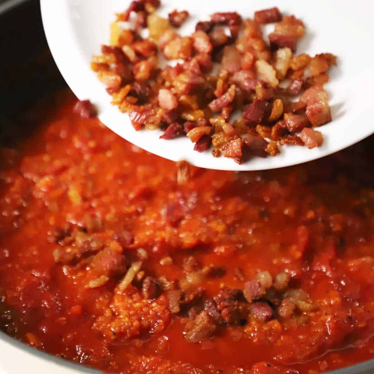 A person dumping cooked guanciale from a white bowl into a skillet filled with a simmering tomato sauce. 