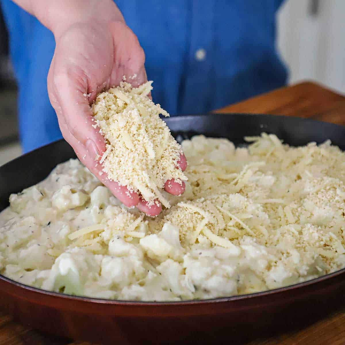 A person sprinkling breadcrumbs mixed with shredded cheese over an unbaked dish of cauliflower gratin. 
