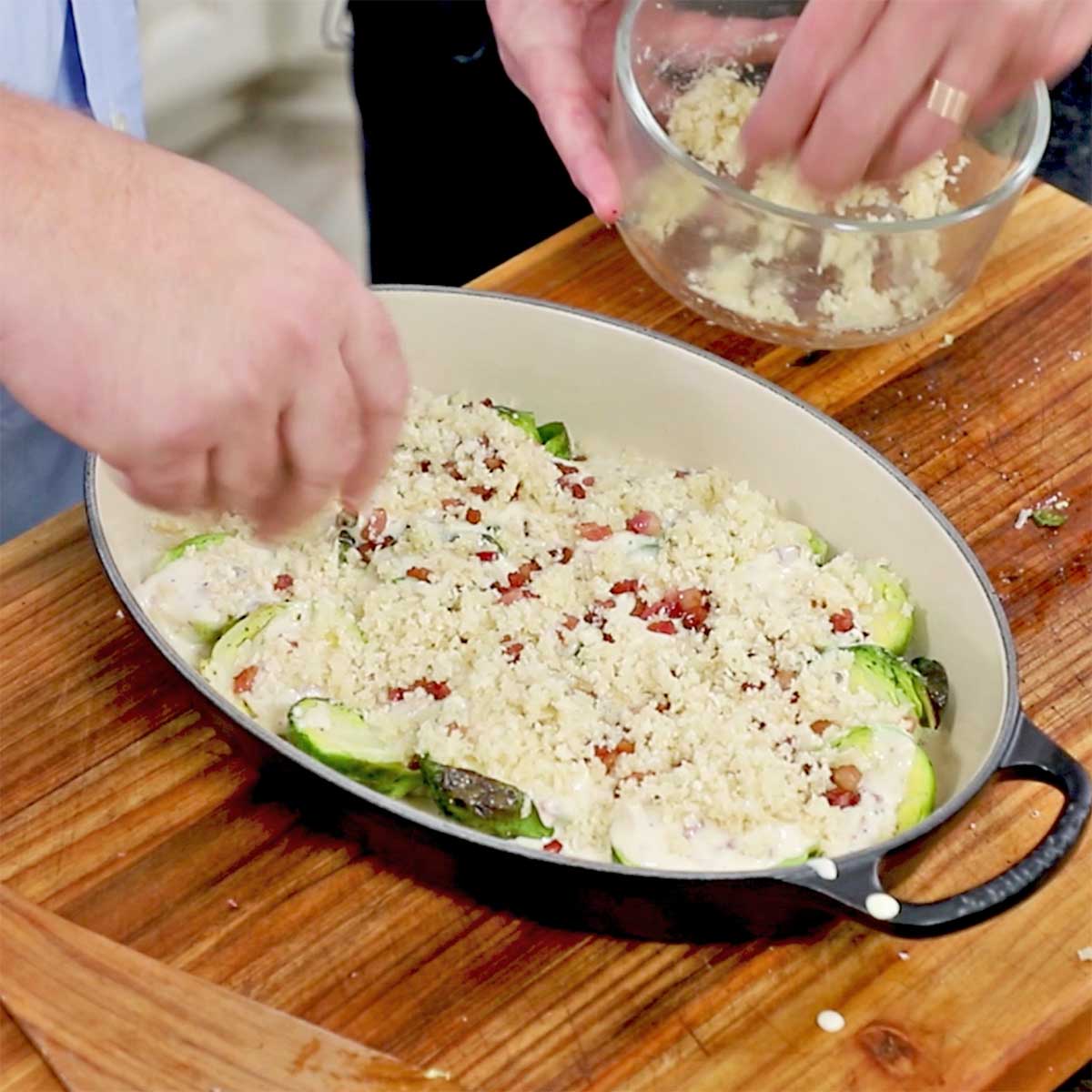 A person sprinkling breadcrumbs over the top of a baking dish the is filled with unbaked Brussels sprouts gratin. 