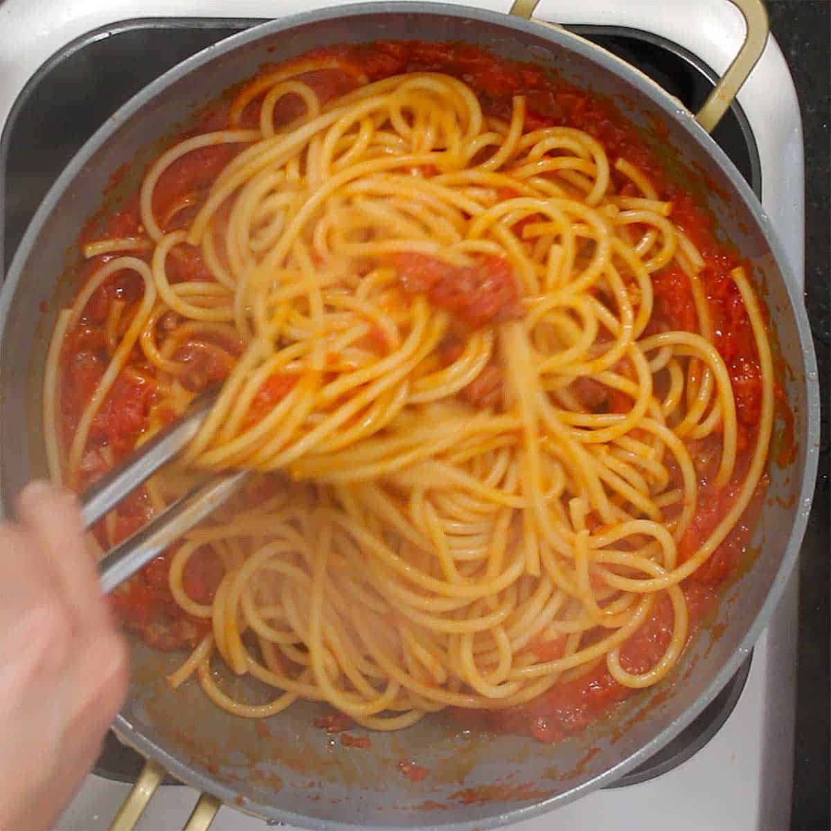 A person using a pair of metal tongs to toss cooked pasta into a skillet filled with a tomato sauce. 
