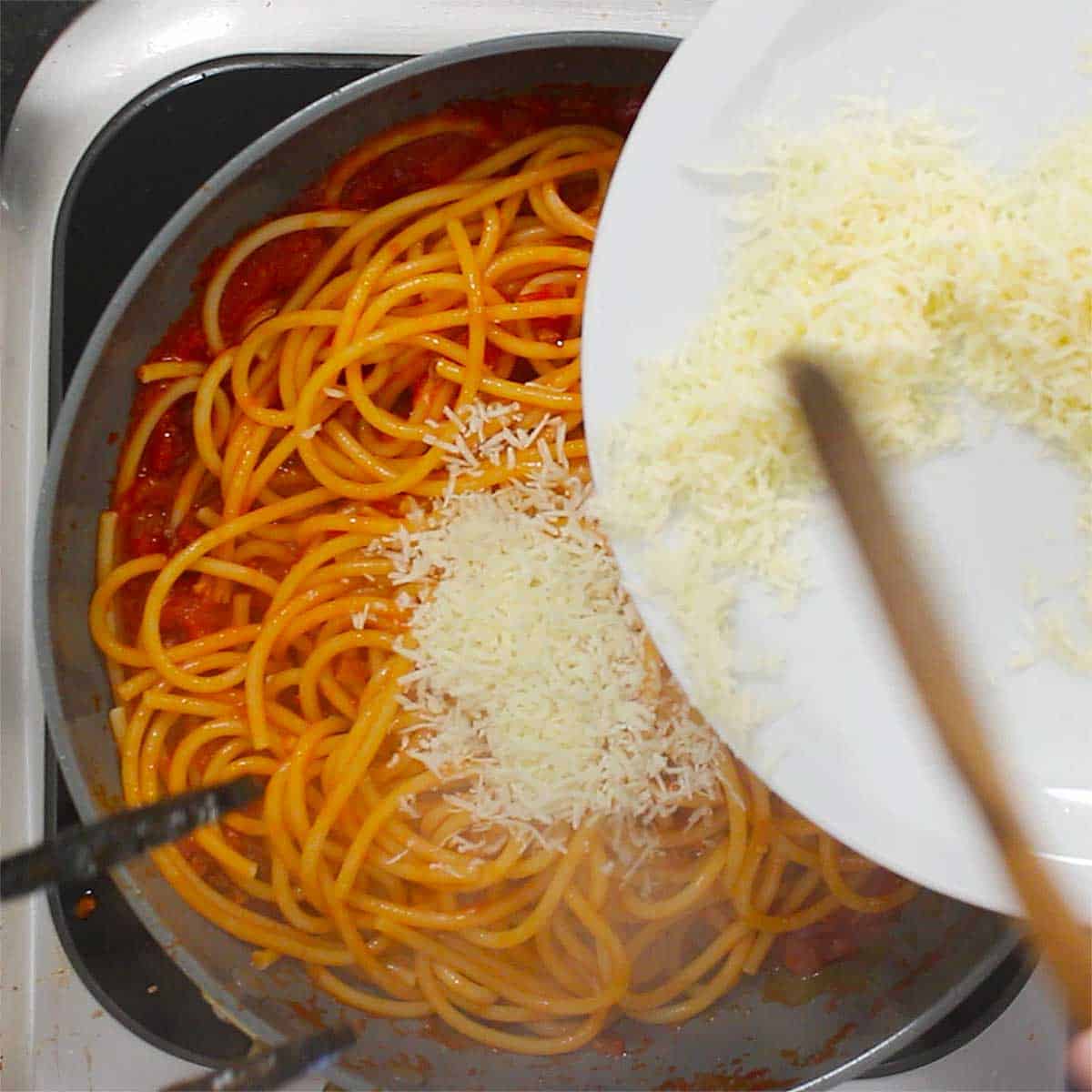 A person using a wooden spatula to transfer grated Pecorino-Romano cheese from a white plate into a skillet filled with pasta in a tomato sauce. 