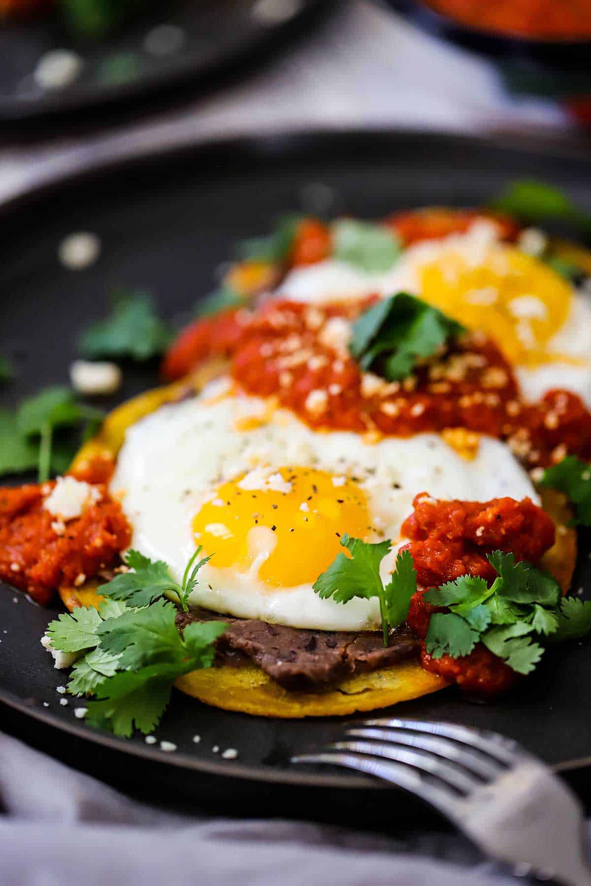 A serving of authentic huevos rancheros on a black dinner plate with a fork nearby. 