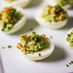 Several rows of avocado deviled eggs resting on a white platter and are garnished with toasted breadcrumbs, bacon, and chives.