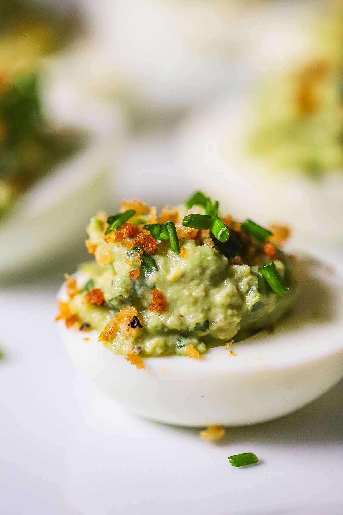 An avocado deviled eggs garnished with toasted breadcrumbs, snipped chives, and crumbled bacon on a white platter. 