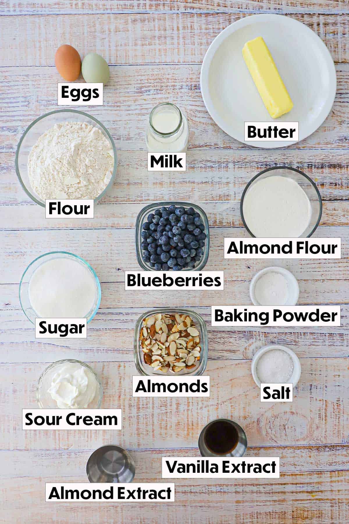The ingredients for blueberry almond coffee cake arranged on a white background, including all-purpose flour, almond flour, fresh blueberries, vanilla extract, almond extract, sugar, butter, eggs, and baking powder, neatly organized for easy preparation.