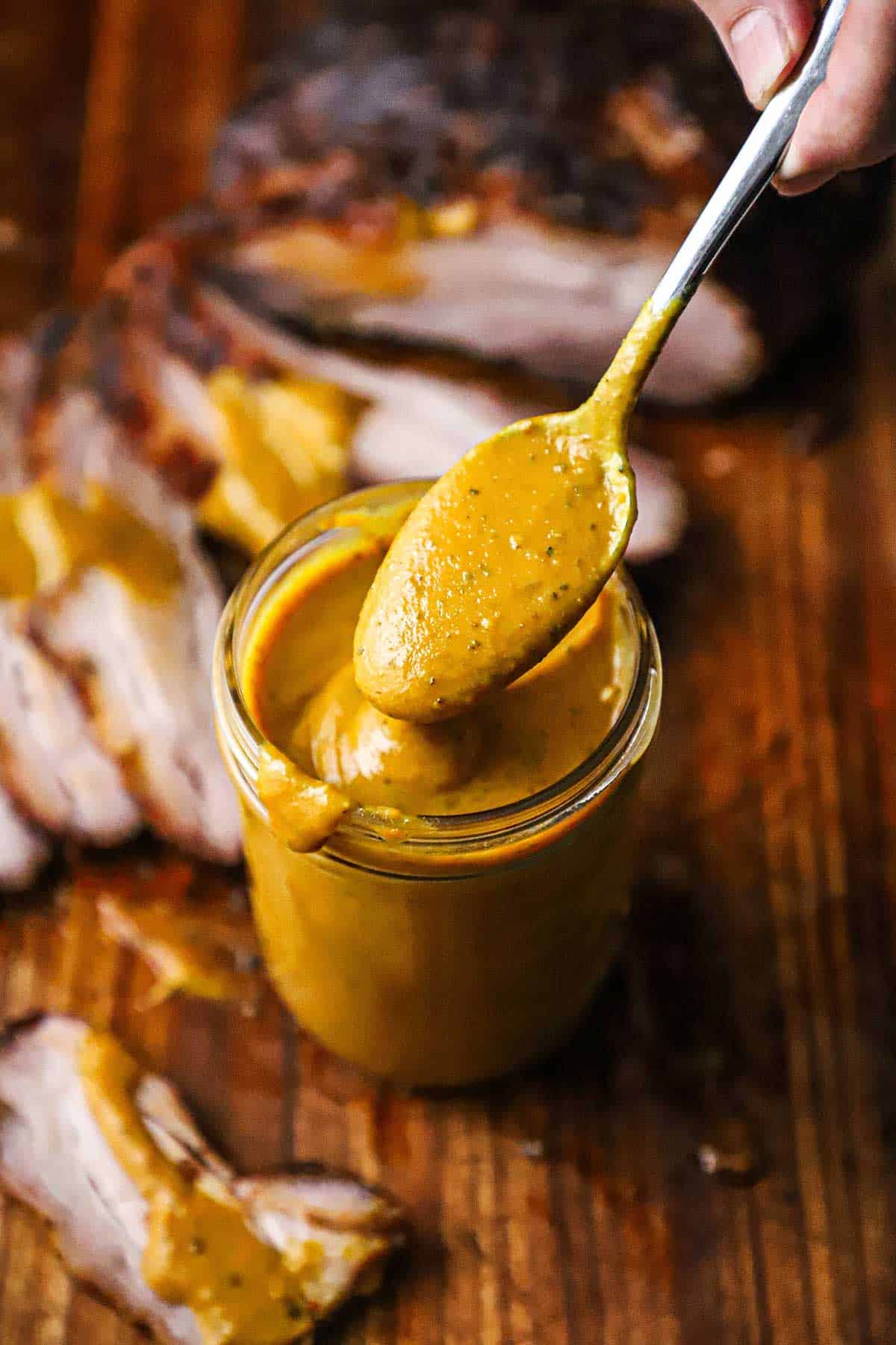 A spoon filled with Carolina mustard sauce being raised from a jar filled with the sauce with a sliced brisket nearby.