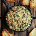 A small cast-iron skillet filled with baked spinach and artichoke dip with several small slices of toasted baguette nearby.