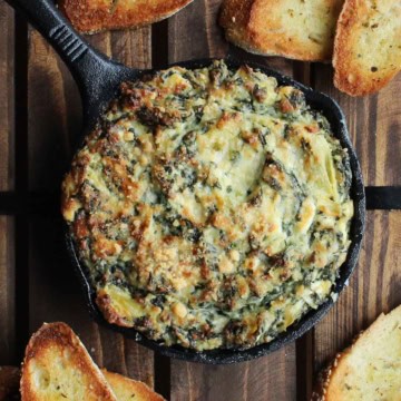A small cast-iron skillet filled with baked spinach and artichoke dip with several small slices of toasted baguette nearby.