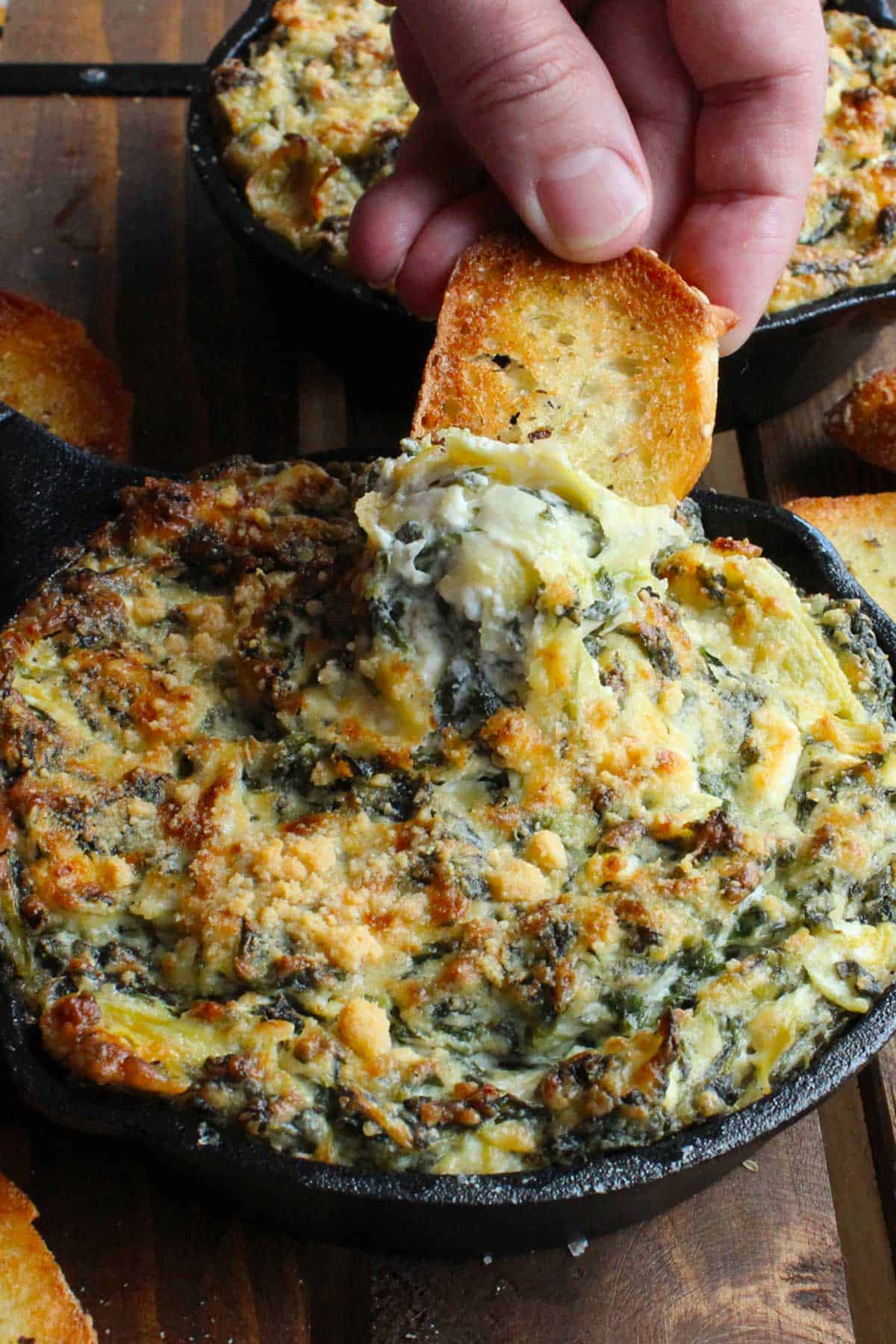 A person lifting a toasted baguette slice out of a cast-iron skillet filled with classic spinach and artichoke dip.