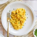 A white plate filled with the fluffiest scrambled eggs that have been garnished with snipped chives and a glass of orange juice nearby.