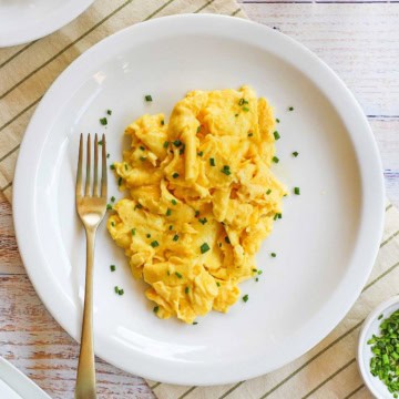 A white plate filled with the fluffiest scrambled eggs that have been garnished with snipped chives and a glass of orange juice nearby.