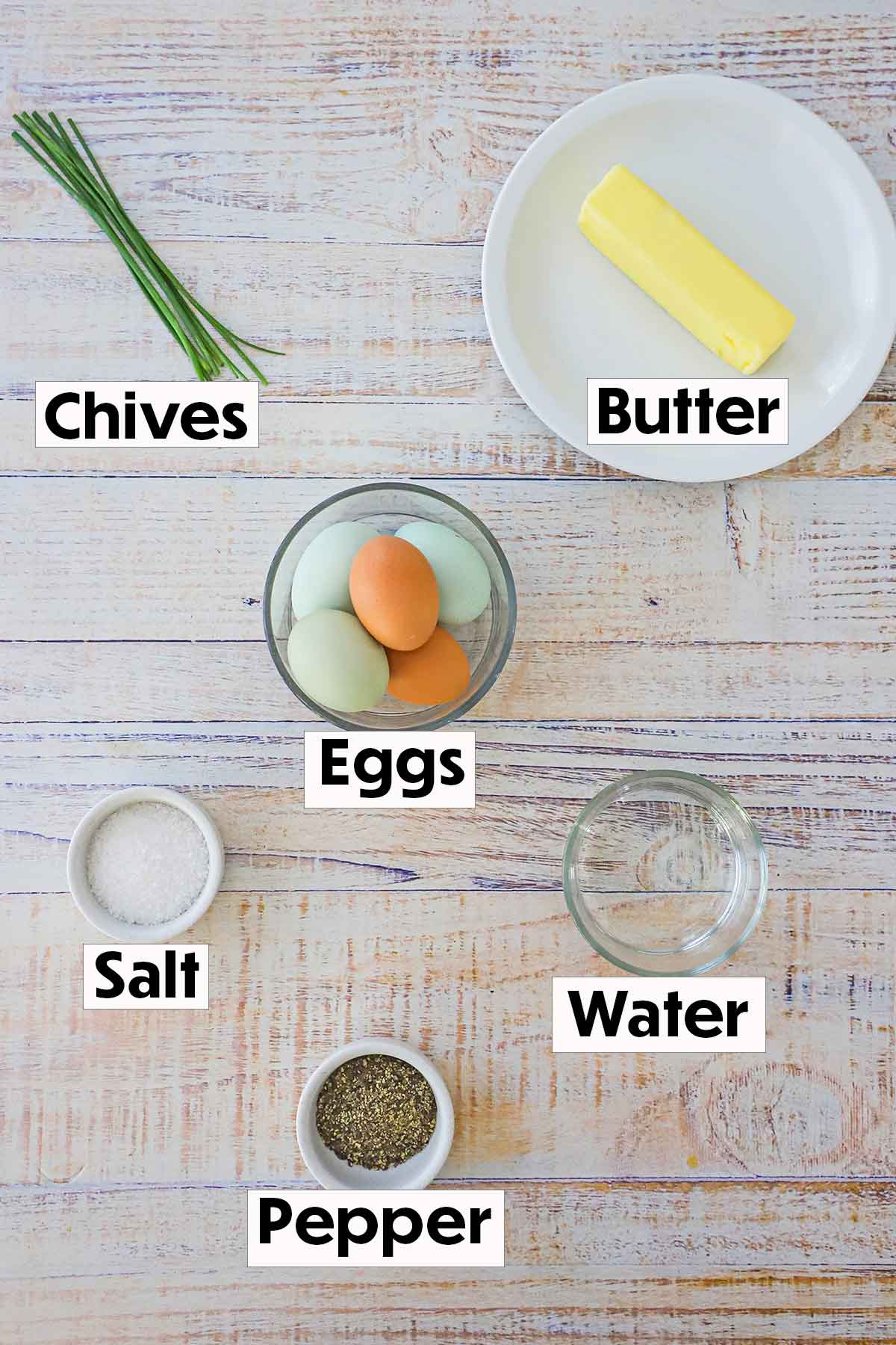 Ingredients for farm-fresh scrambled eggs including eggs, butter, salt, pepper, water, and chives all arranged on a white background. 