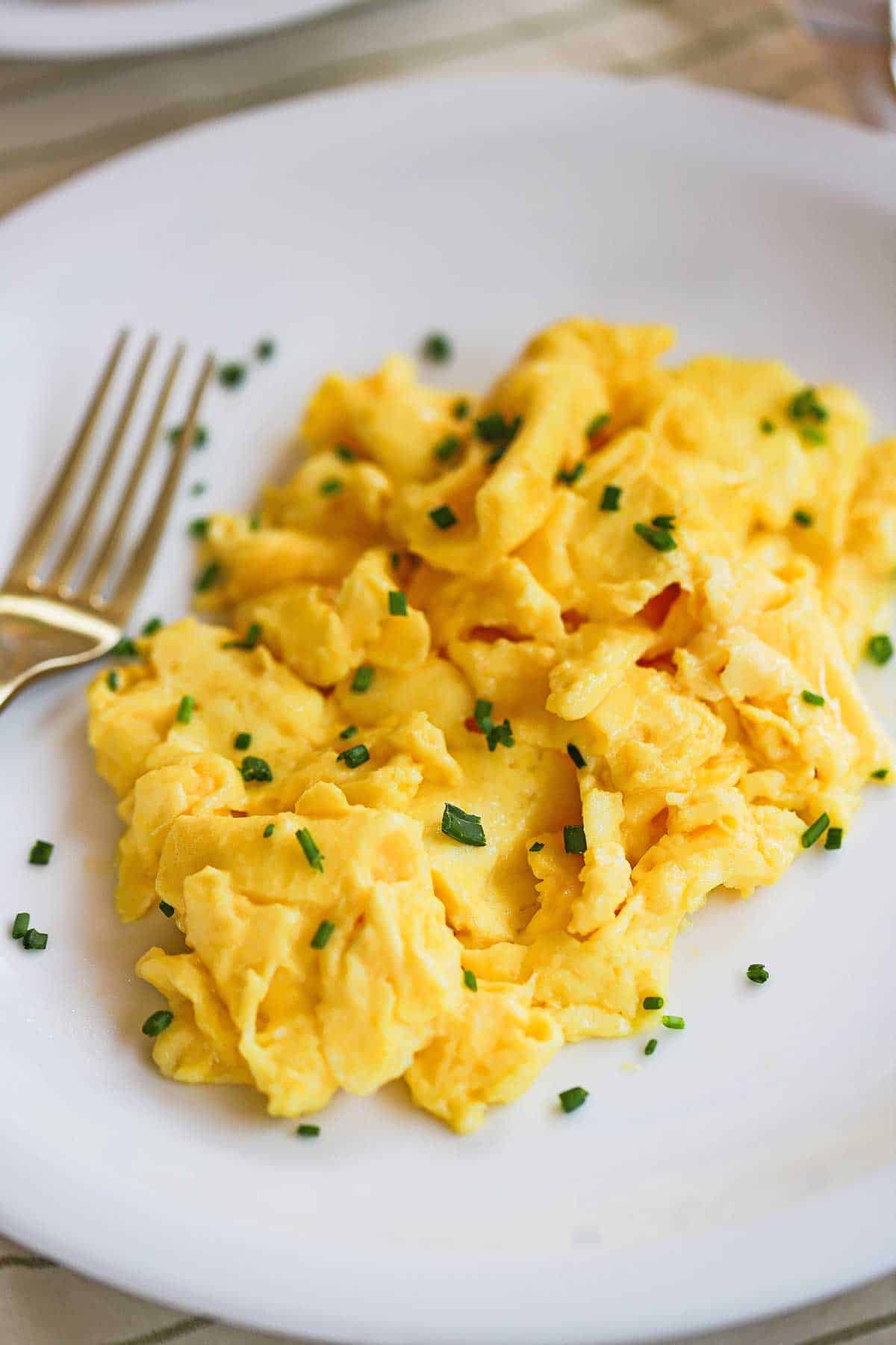 A pile of fluffiest scrambled eggs topped with snipped chives on a white dinner plate with a gold fork sitting next to it. 