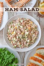 A shallow white bowl filled with best-ever ham salad surrounded by a plate of green leaf lettuces, slices of bread, plates, and glasses of iced tea.