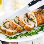 A sliced herb-stuffed pork loin on a white platter with fresh herbs and a chef's knife on the platter next to the roast.