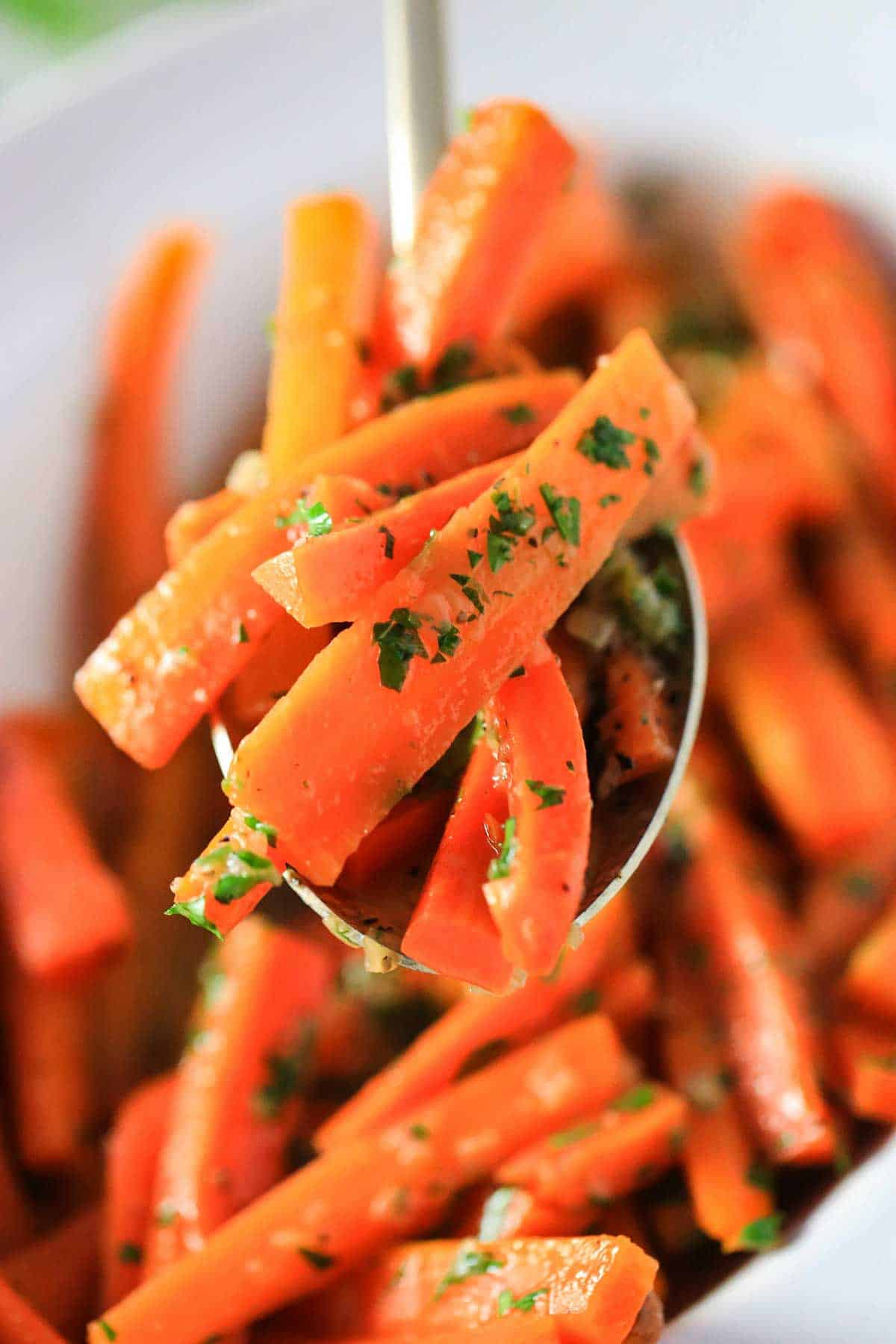 A large serving spoon raising a serving of maple-glazed carrots over a bowl of the same thing.