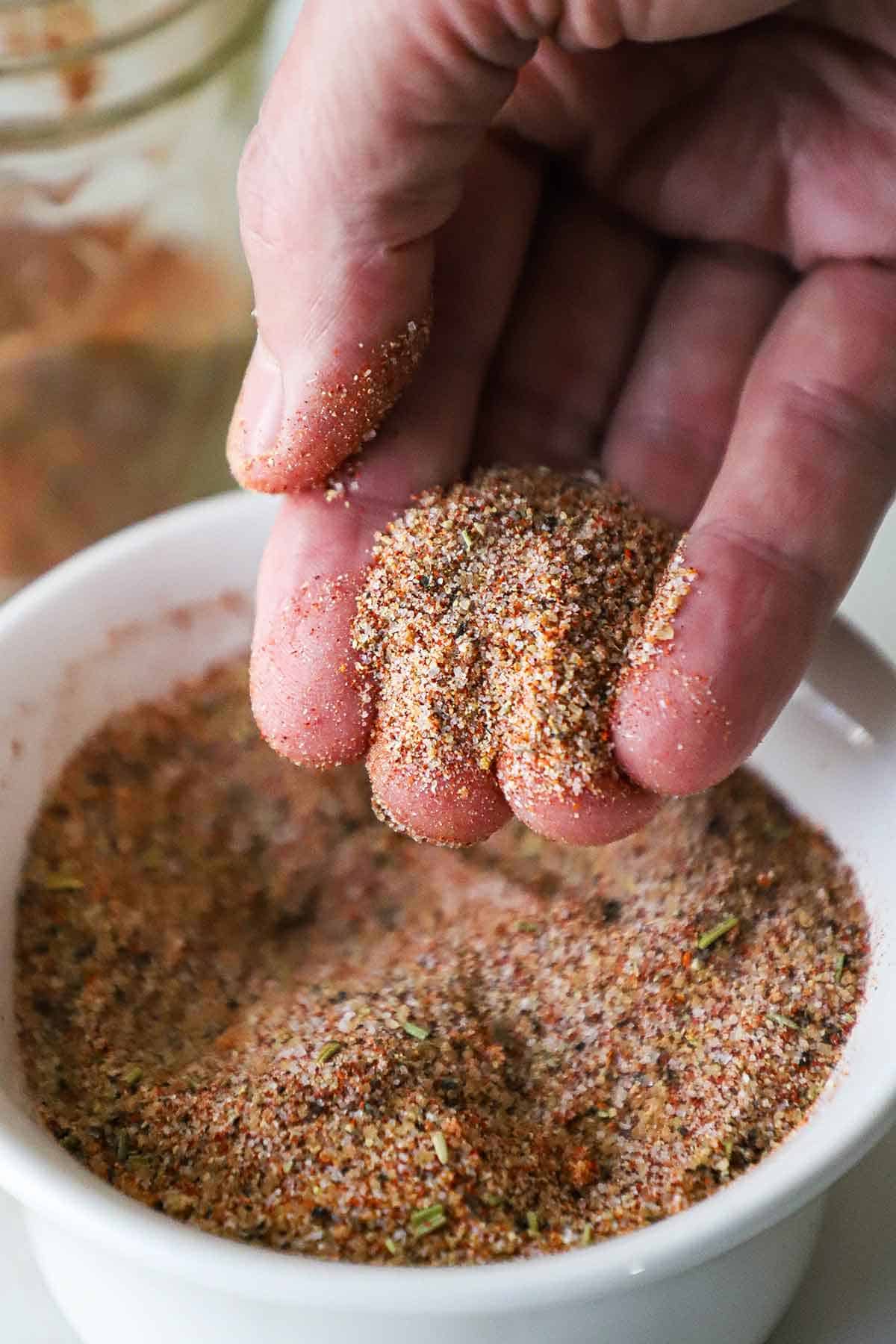 A person holding a small pile of pork rub in his fingers over a bowl of the rub.