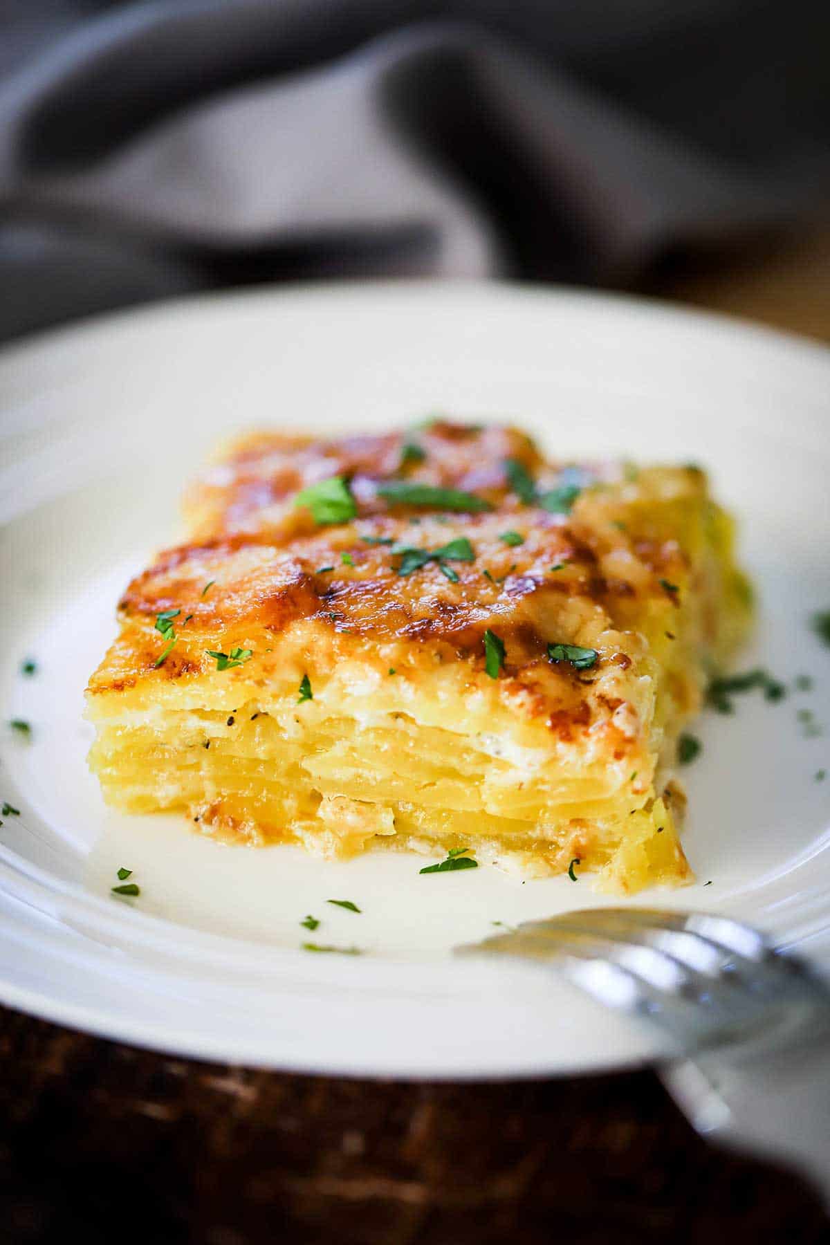 A square slice of potatoes dauphinoise on a white plate and is garnished with chopped Italian parsley.