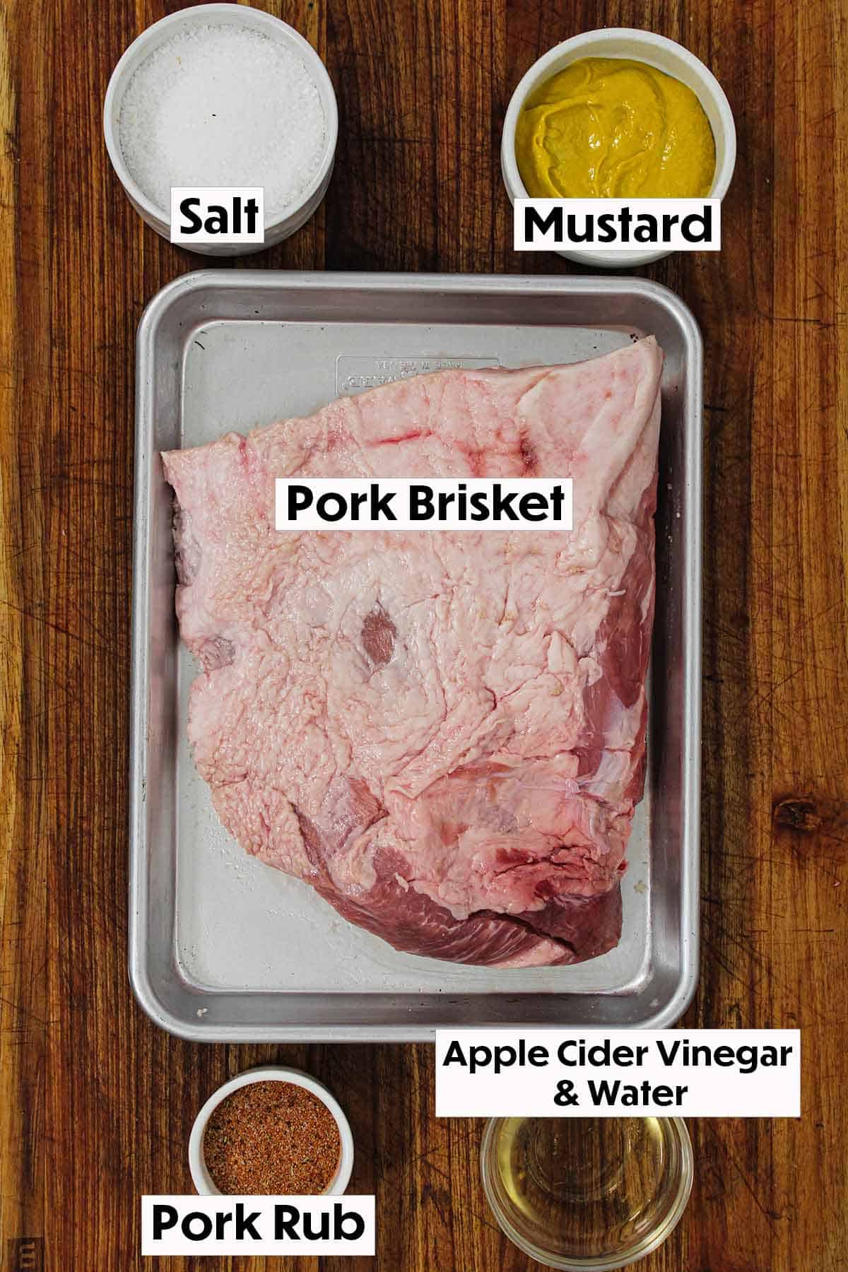 An arrangement of ingredients on a wooden cutting board for smoked pork brisket including an uncooked pork brisket, salt, yellow mustard, pork rub, and apple cider vinegar. 