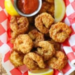A plastic basket lined with a red and white checkered wax paper and filled with Southern-fried shrimp, a couple of lemon wedges, and a small vessel of homemade cocktail sauce.