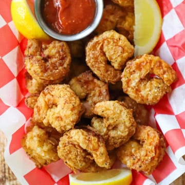 A plastic basket lined with a red and white checkered wax paper and filled with Southern-fried shrimp, a couple of lemon wedges, and a small vessel of homemade cocktail sauce.