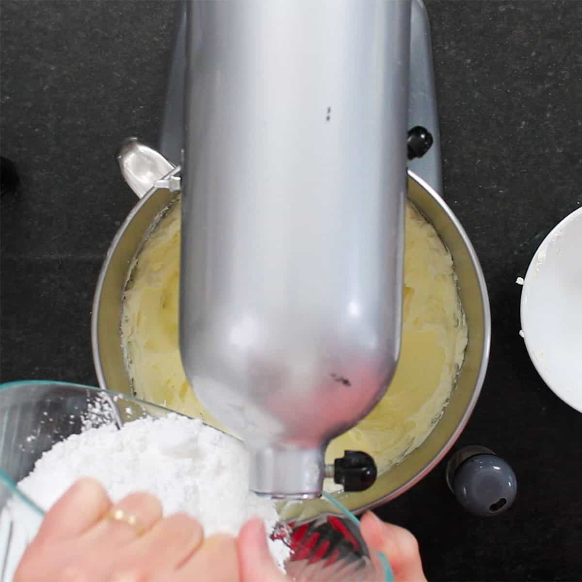 A person adding powdered sugar into the bowl of a stand mixer that is filled with creamed butter and cream cheese. 