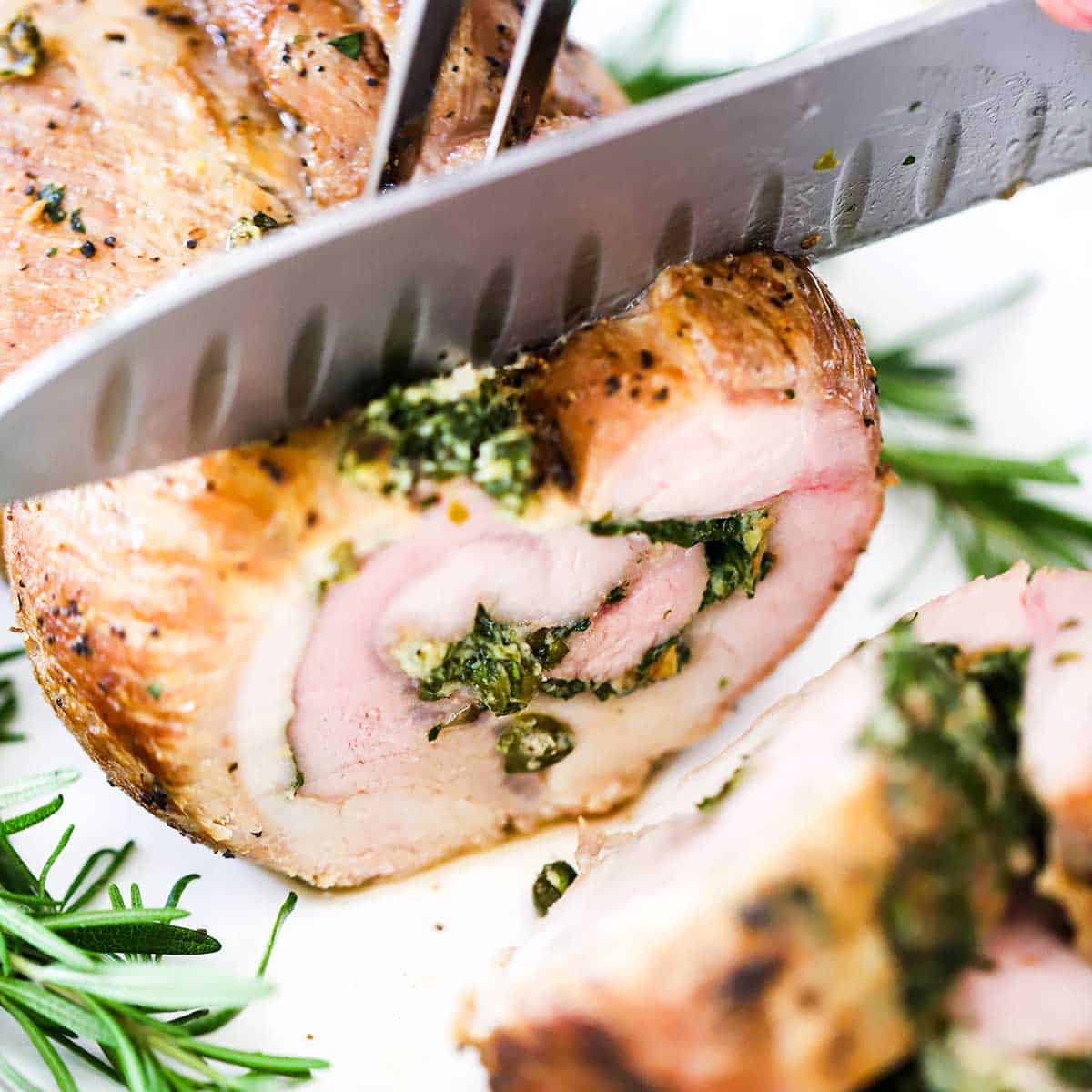 A person using a large chef's knife to slice an herb-stuffed pork loin on a white platter. 