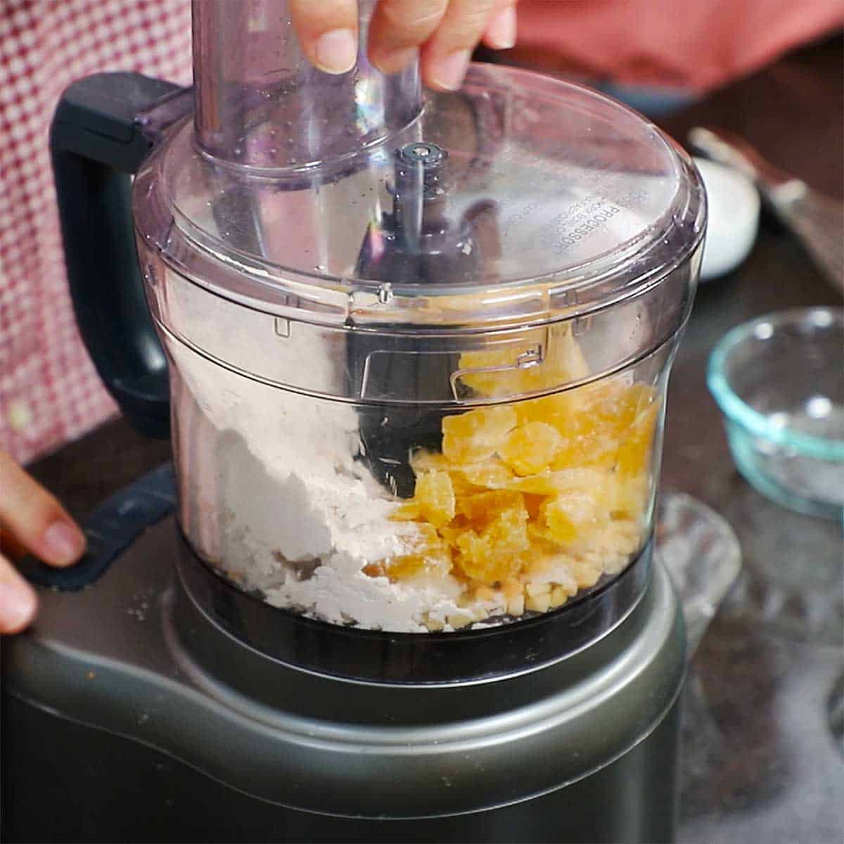 A person using a food processor to chop crystalized ginger, flour, macadamia nuts, and coconut. 