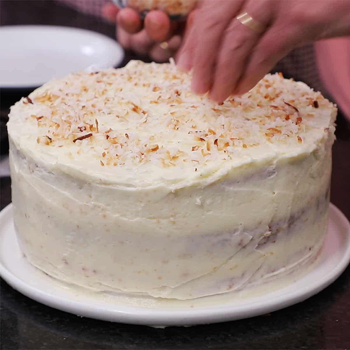 A person sprinkling toasted coconut over the top of a frosted three-layer carrot cake. 