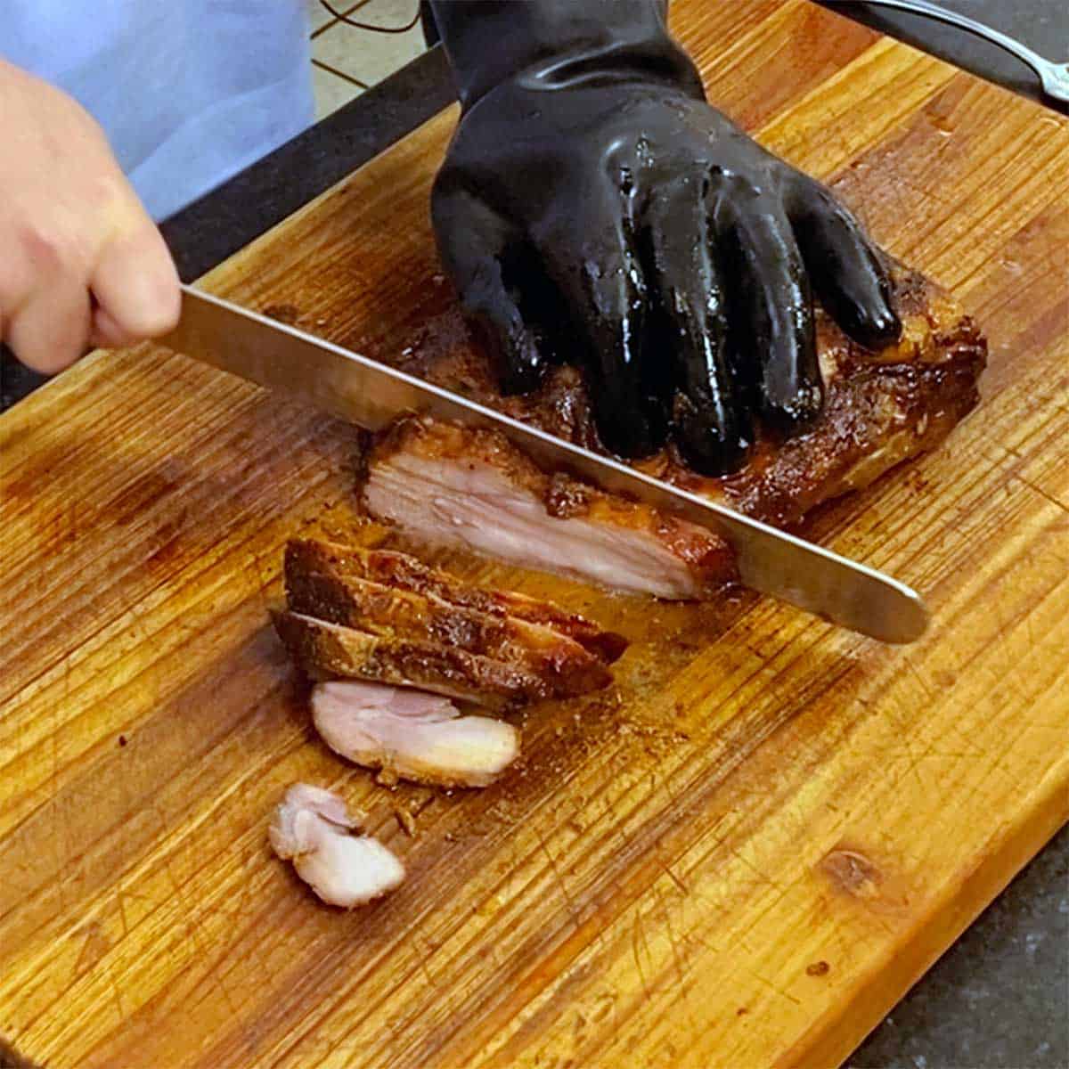 A person wearing a heat-resistant glove on one hand using a large knife in the other hand to slice a smoked pork brisket. 