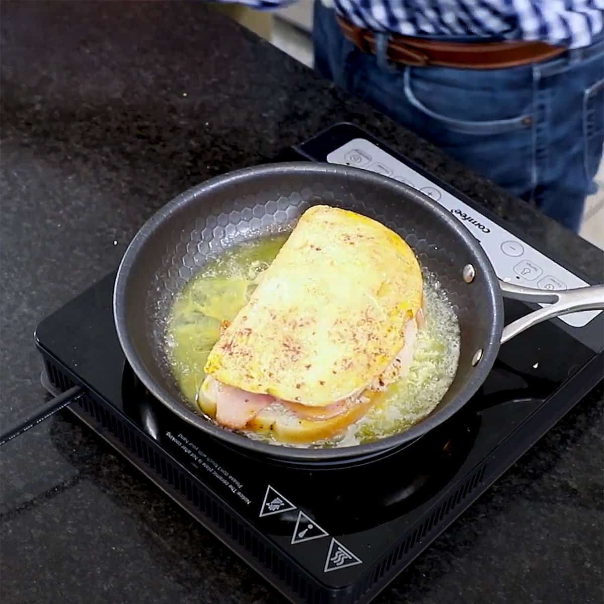 A Monte Cristo sandwich being cooked and lightly fried in a skillet filled with simmering melted butter on a stovetop. 