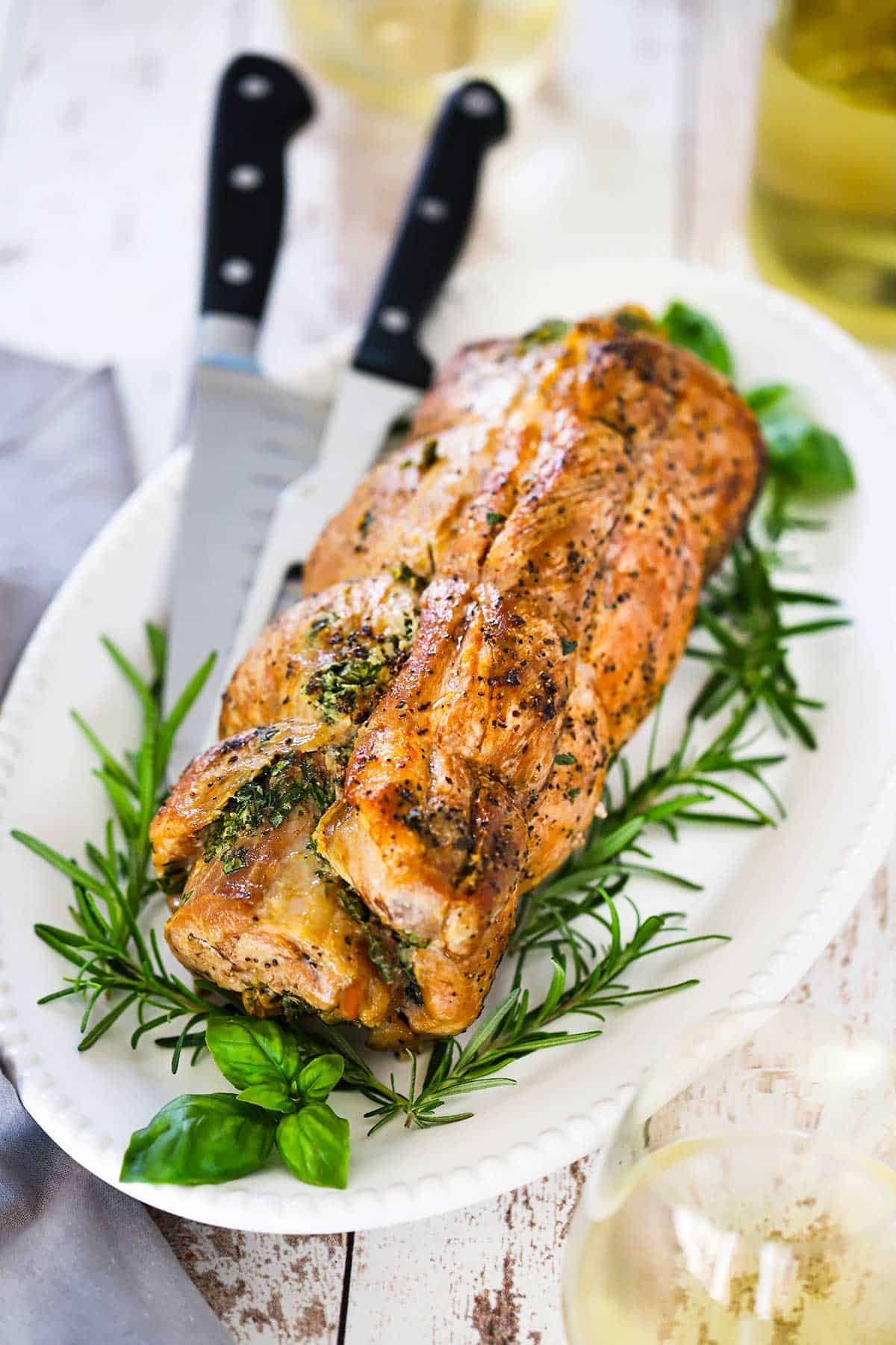 A fully cooked herb-stuffed pork loin resting on a white platter with fresh rosemary on each side of it. 