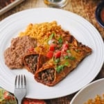 A white plate filled with two beef enchiladas with red sauce next to a serving of Mexican rice and refried beans.