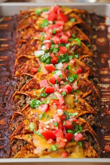 A sheet pan filled with a row of beef enchiladas with red sauce and topped with chopped tomatoes and lettuce.