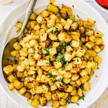 A white bowl filled with easy breakfast potatoes garnished with chopped parsley with a large serving spoon inserted along the side of the dish.