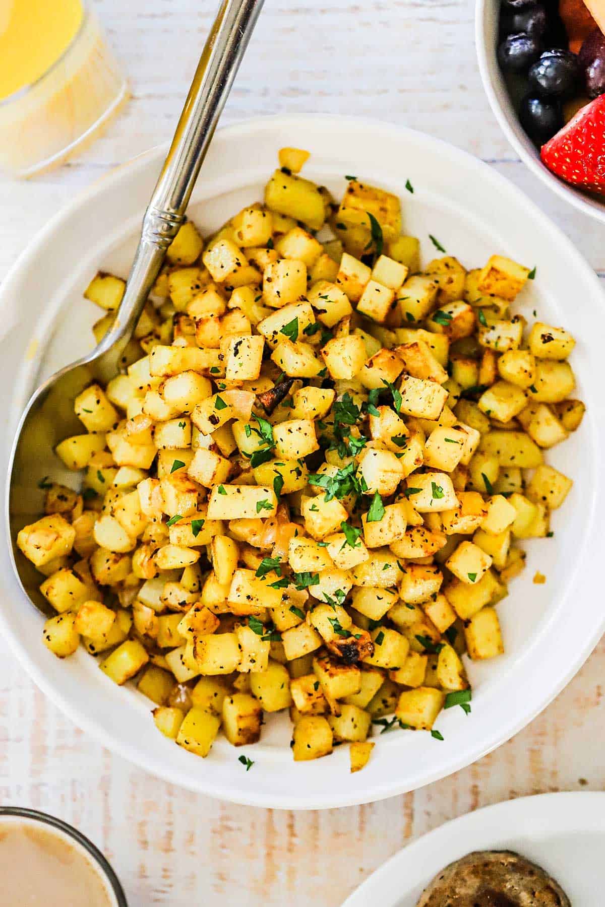 A white bowl filled with easy breakfast potatoes garnished with chopped parsley with a large serving spoon inserted along the side of the dish.