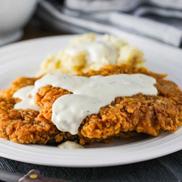 A crispy Southern chicken fried steak on a white dinner plate topped with cream gravy and next to a pile of mashed potatoes.