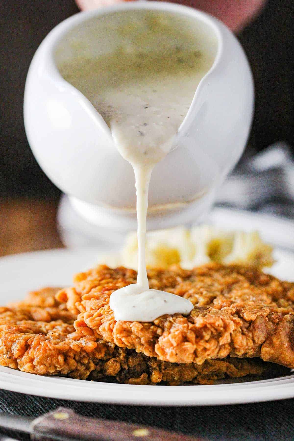 A person pouring cream gravy from a gravy boat over the top of Southern chicken fried steak on a white dinner plate. 