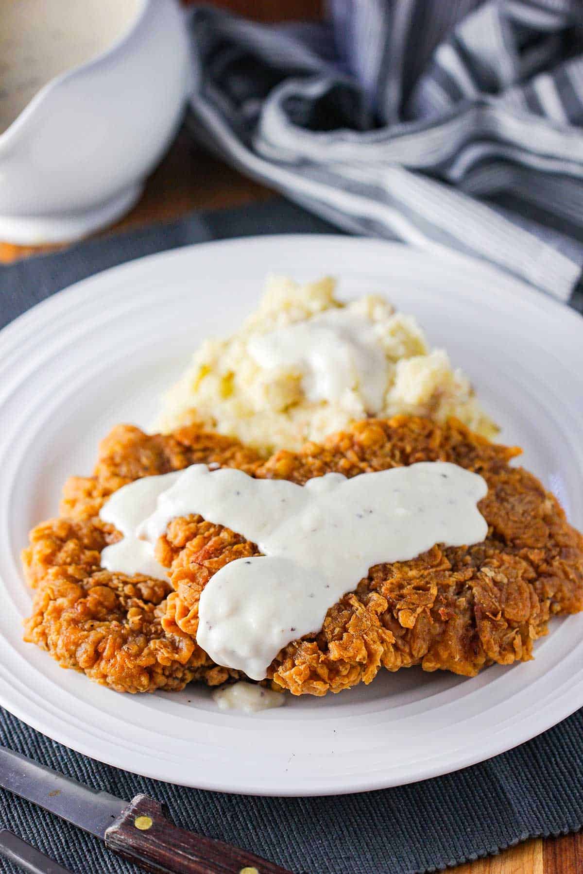 A crispy Southern chicken fried steak on a white dinner plate topped with cream gravy and next to a pile of mashed potatoes.