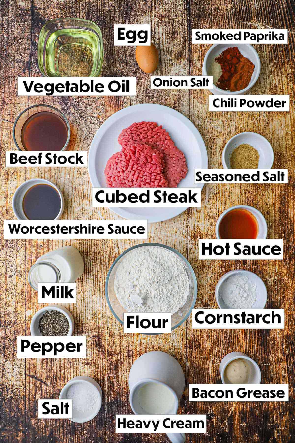 The ingredients for chicken fried steak with cream gravy arranged on a wooden background, including tenderized beef steak, flour, eggs, milk, heavy cream, seasonings, and a small bowl of cream for the gravy, all neatly organized for easy preparation.