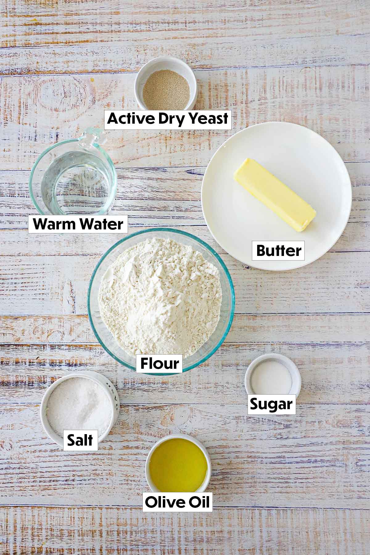Ingredients for homemade dinner rolls displayed on a white background, including all-purpose flour, yeast, sugar, salt, milk, butter, and eggs, arranged neatly for easy preparation.
