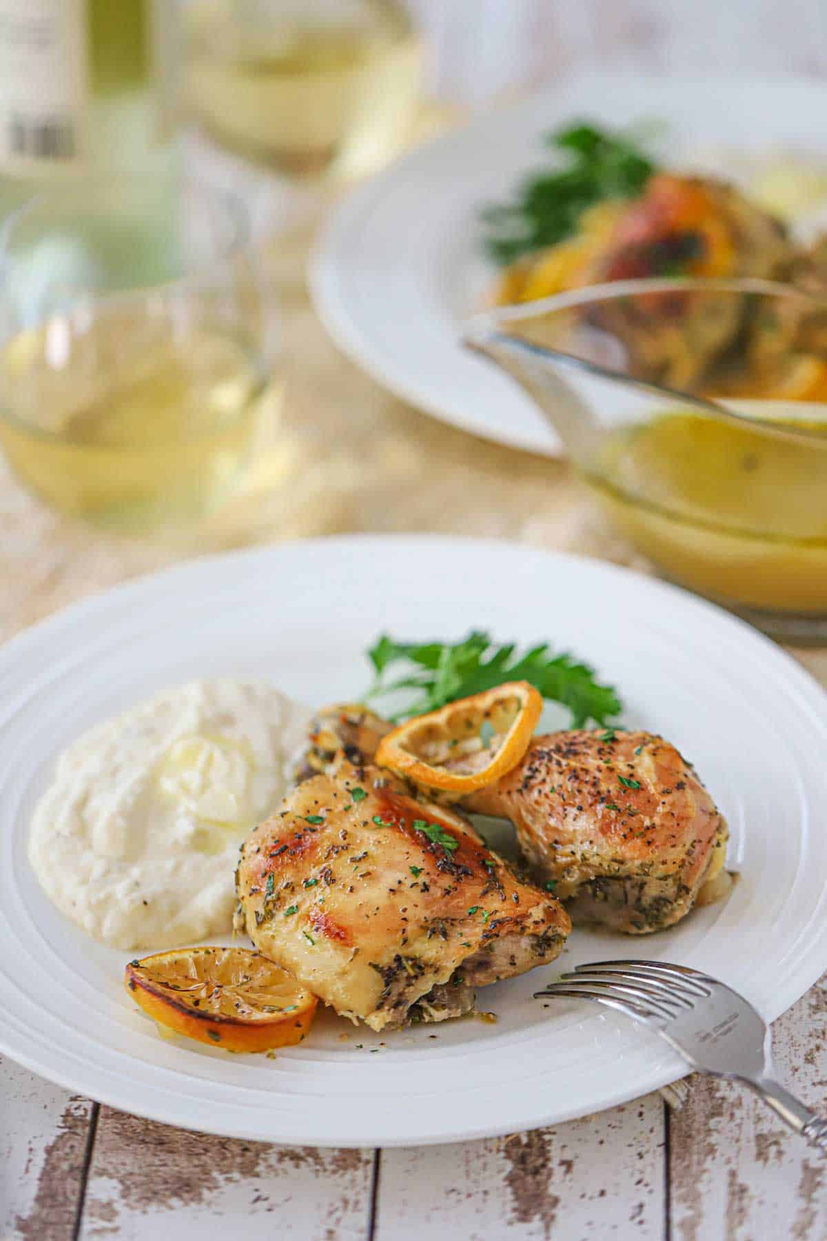 A white dinner plate filled with two pieces of lemon roast chicken sitting next to a pile mashed potatoes, grilled lemon slices, and fresh parsley.