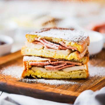 Powdered sugar being sprinkled over the tops of a Monte Cristo sandwich that has been cut in half and placed on top of each other.