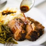 A person pouring an au jus sauce from a spoon onto slices of slow-cooker brisket on a white plate next to a pile of green beans and mashed potatoes.